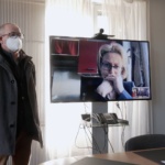 Dans un bureau, un grand écran plat affiche l'image d'une quinquagénaire aux traits du visage préoccupés. A côté de cet écran se tient un homme masqué.