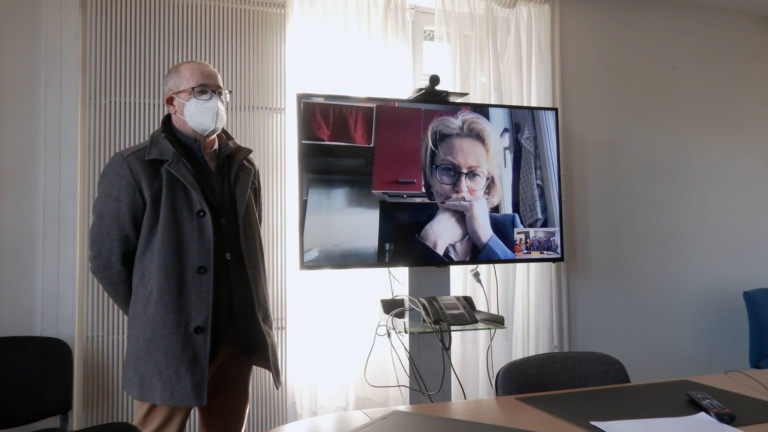 Dans un bureau, un grand écran plat affiche l'image d'une quinquagénaire aux traits du visage préoccupés. A côté de cet écran se tient un homme masqué.