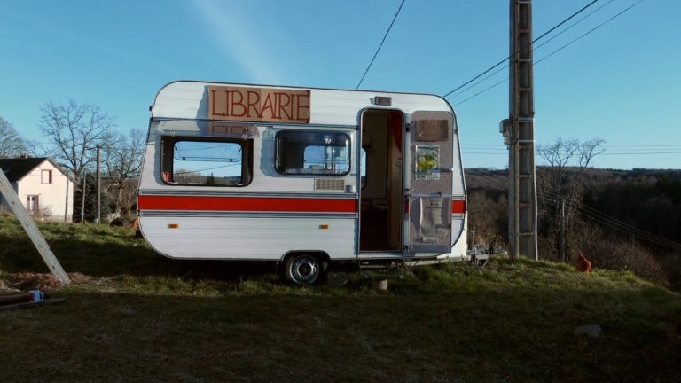 Une caravane blanche et rouge trône sur une pelouse en pleine campagne. Sur les rebords de ses fenêtres ouvertes est posé un carton où il est écrit en grosses lettres le mot "Librairie". Une poule picore à côté de la caravane.