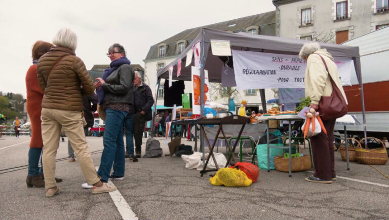 Un petit groupe de personnes échangent devant un stand déployé sur un parking pavé. Sur une banderole, on peut lire "Sans papiers, régularisation durable pour toutes".