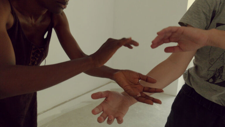 Dans une pièce aux murs nus, deux personnes se font face. Elles tendent leurs mains l'une vers l'autre, paume ouverte, sans qu'elles ne se touchent.