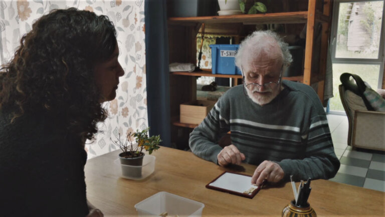 Un vieil homme aux cheveux et à la barbe blanche est attablé dans un salon. Il manipule avec attention des lettres de Scrabble posés sur une petite surface blanche encadrée. Face à lui, une quadragénaire aux longs cheveux bouclés observe ses gestes.