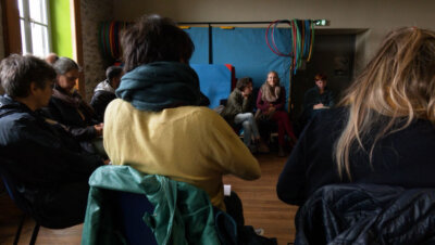 Un groupe d'adultes sur assis en cercle dans une large pièce. Elles s'écoutent attentivement.