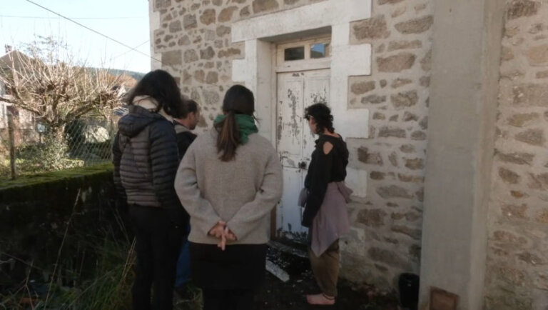 Quatre personnes observent depuis un jardin la porte d'entrée d'une maison de campagne. Le battant de la porte en bois est marqué par des traces d'écaillement de peinture et de coups. Des éclats de peinture sont parsemés au sol.