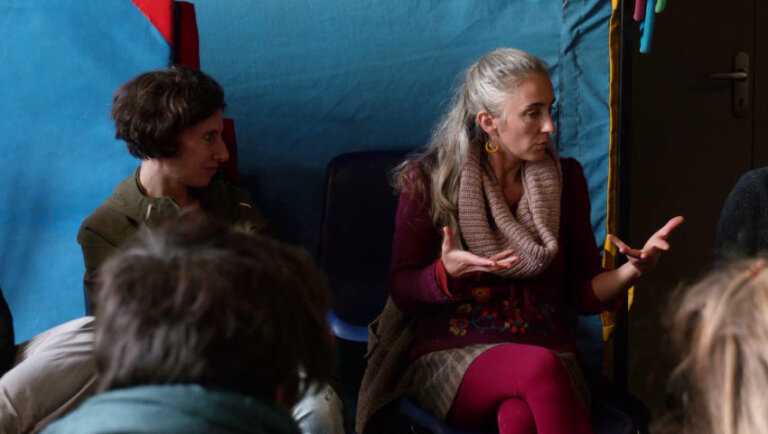 Dans une pièce, un groupe de personnes sont assises sur des chaises disposées en cercle. Sous le regard attentif d'une femme brune avec une coupe au carré, une femme aux longs cheveux argentés s'adresse au groupe. Elle appuie ses propos d'un geste de ses mains, dont les paumes sont tournées vers le haut.