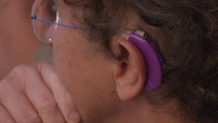 Une femme aux cheveux courts bouclés affiche un air concentré, sa main posée sur le menton. Elle porte à son oreille un implant auditif mauve.