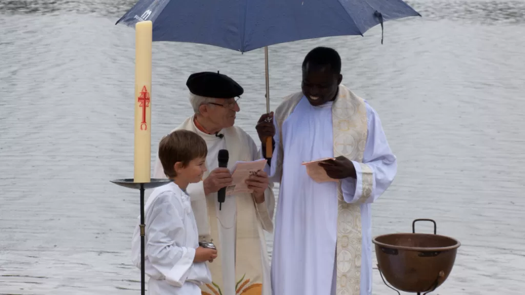 Deux prêtres et un enfant, vêtus de blanc, dehors sous un parapluie. Autour d'eux, un grand cierge et un bénitier métallique. L'un des prêtres est âgé et blanc, l'autre jeune et noir. Derrière eux, une étendue d'eau sous la pluie.