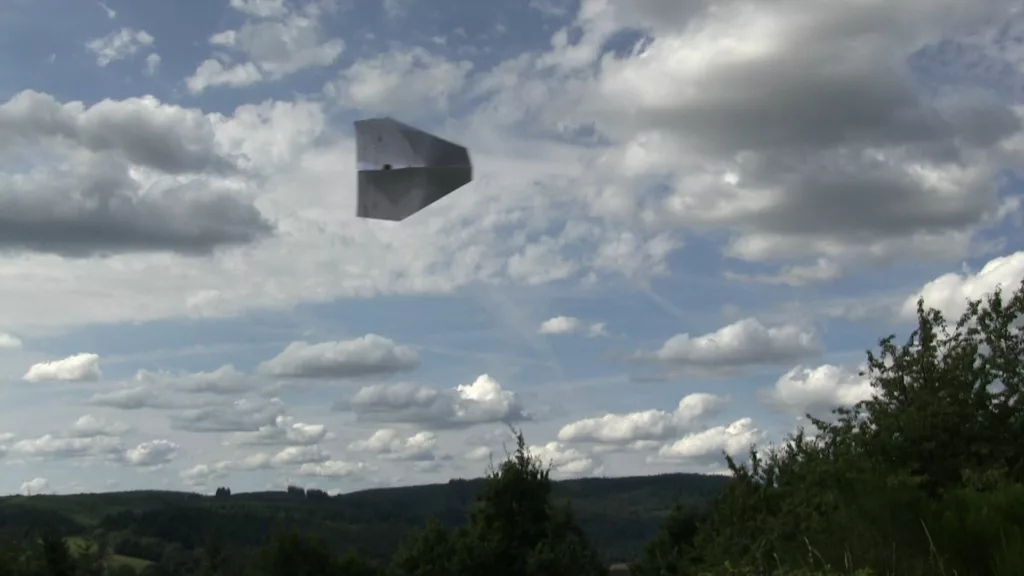 Un avion en papier vole devant un ciel parsemé de nuages blancs, au-dessus de collines forestières.