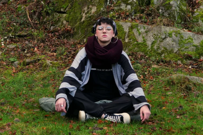 Une jeune femme portant un châle se tient en tailleur sur l'herbe. Elle ferme les yeux, l'air calme.