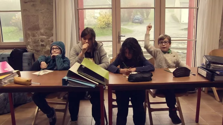 Quatre enfants sont assis dans une salle, face à des tables encombrées de classeurs et cahiers. L'un d'eux lève le doigt avec enthousiasme tandis que ses camarades sont plus ou moins avachis.
