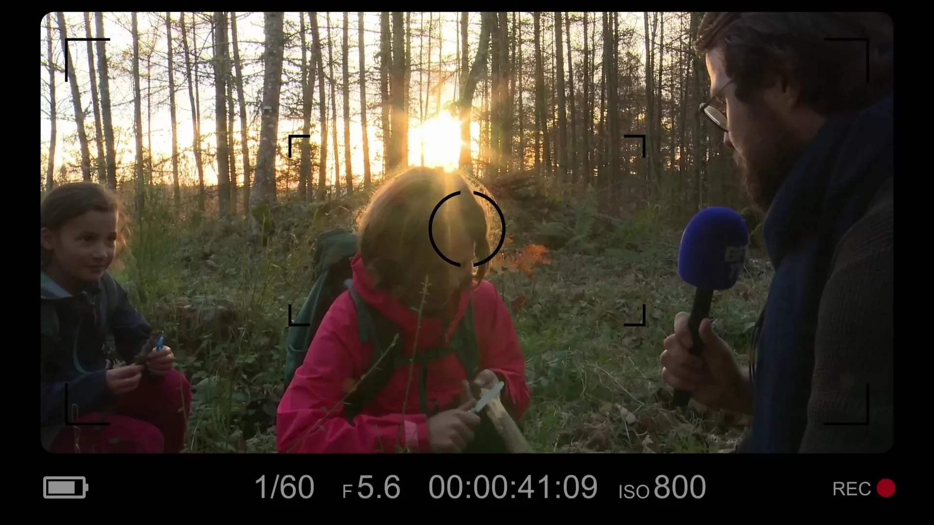 Un journaliste interviewe deux jeunes filles dans un bois au coucher du soleil.