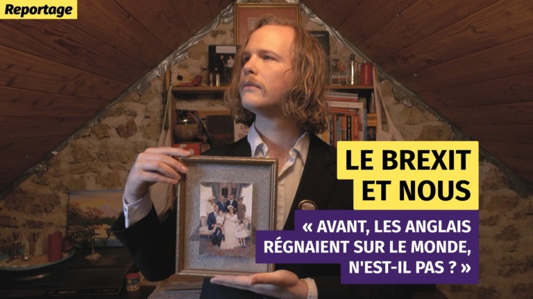Portrait en plan buste d'un homme vêtu d'un costume noir, la trentaine, avec une vieille photo de famille encadrée, soigneusement tenue entre ses mains. Intérieur, grenier aménagé.