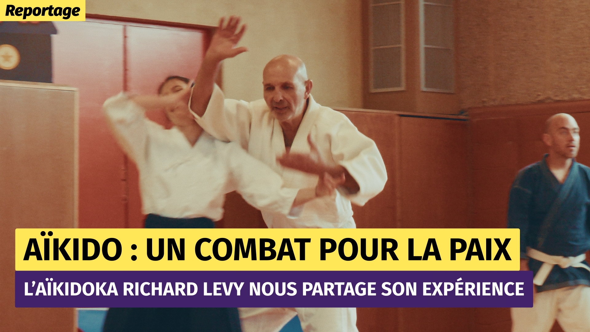 À l'intérieur d'un gymnase, on aperçoit deux personnes vêtues de kimonos blancs. Un homme d'un certain âge, chauve, a les bras levés de manière théâtrale et bouscule l'autre personne.